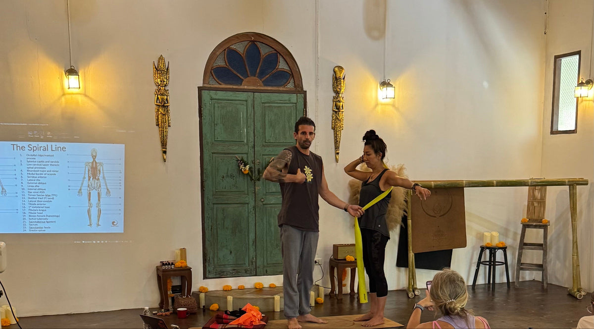 A group of people practicing yoga in a spacious indoor studio with a concrete floor, hanging lantern lights, and a green wooden door in the background. The teacher is demonstrating the spiral line using a resistance band. 